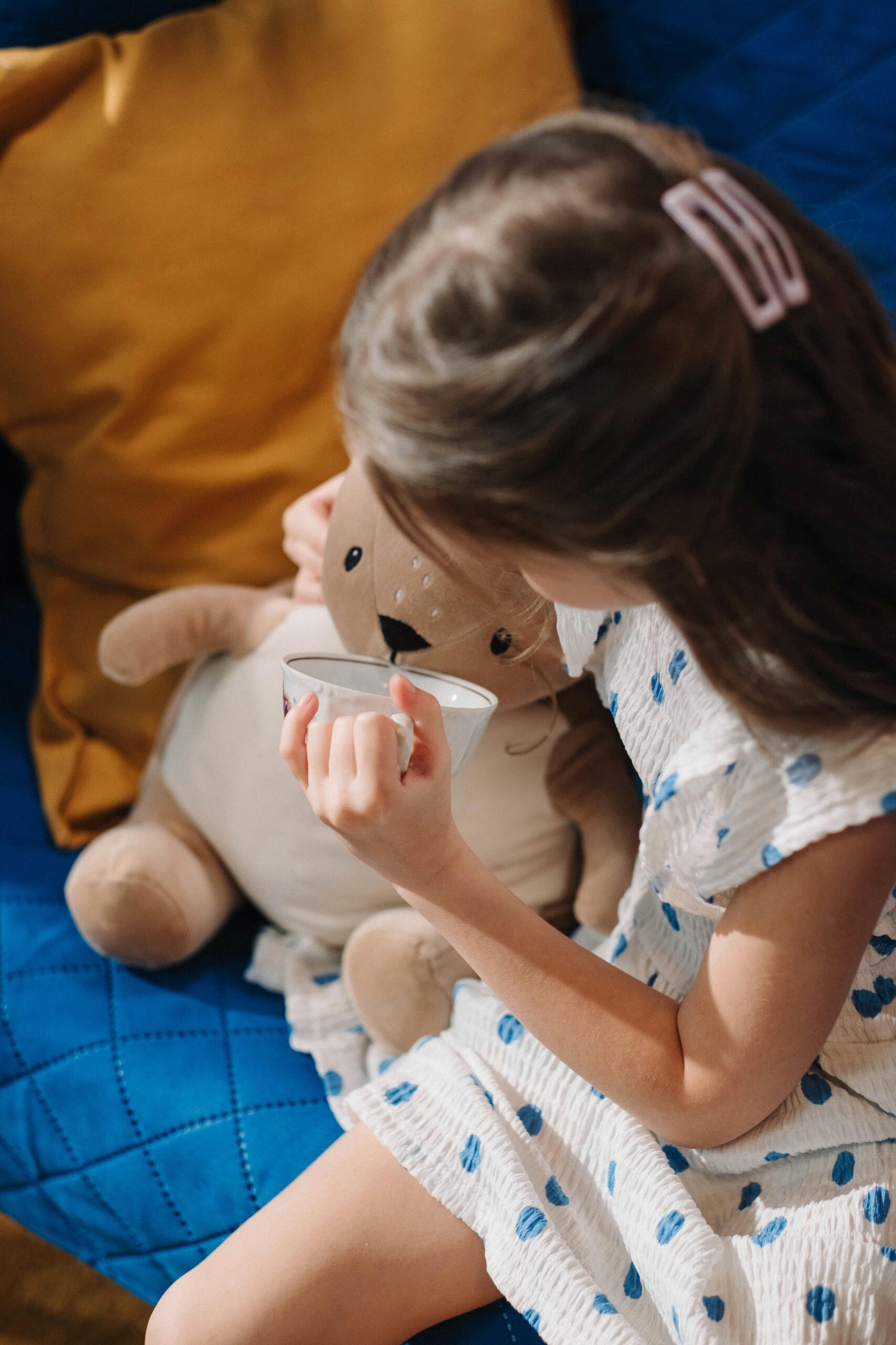 A young girl in a polka-dot dress enjoys a drink with her stuffed toy friend indoors.